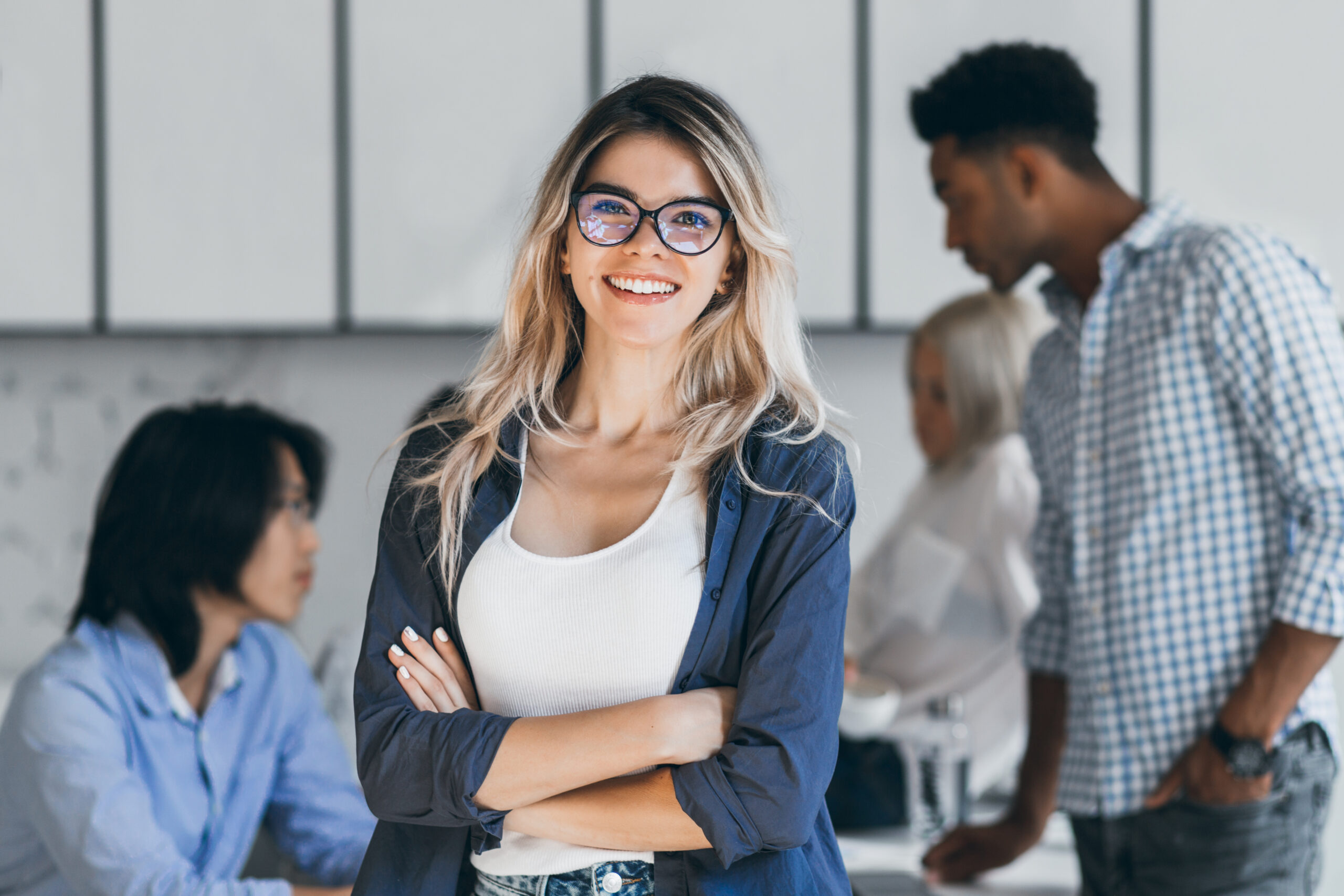 Confident blonde female manager posing with smile after conference with other employees. Asian programmer talking with african freelancer while fair-haired secretary laughing on foreground..